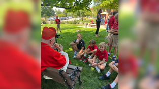 Elderly man in red robe talks to kids in uniforms sitting on grass. Sunny park setting, trees and people in background. Peaceful mood.