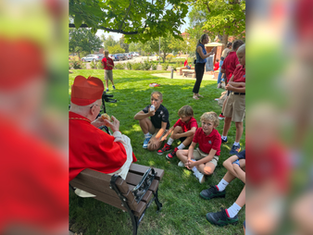 Elderly man in red robe talks to kids in uniforms sitting on grass. Sunny park setting, trees and people in background. Peaceful mood.