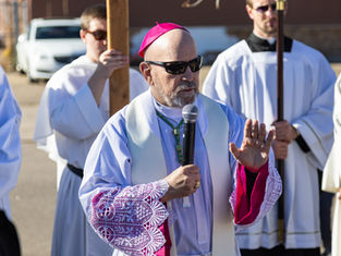 A priest in a magenta hat and lace sleeves holds a microphone, speaking outdoors. Others in white robes stand behind him, one holding a cross.