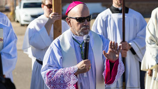 A priest in a magenta hat and lace sleeves holds a microphone, speaking outdoors. Others in white robes stand behind him, one holding a cross.