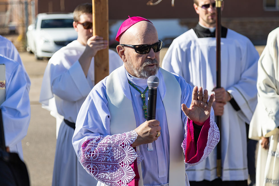 A bishop wearing a pink cap and sunglasses speaks into a microphone outdoors, with priests in white garments holding crosses behind him.