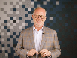 Smiling man in glasses and plaid jacket stands against a patterned pixelated background, conveying a cheerful mood.