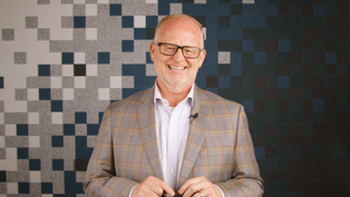 Smiling man in glasses and plaid jacket stands against a patterned pixelated background, conveying a cheerful mood.