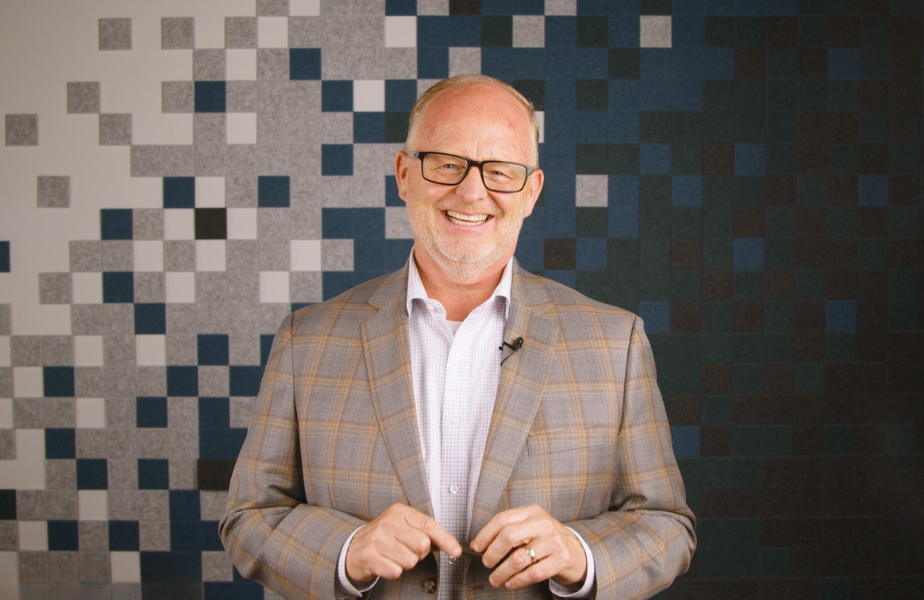 Smiling man in glasses and plaid jacket stands against a patterned pixelated background, conveying a cheerful mood.