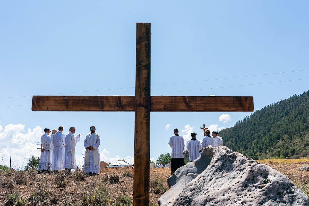 White-robed priests gather outdoors around a large wooden cross, with mountains and blue sky behind them, in a solemn ceremony.