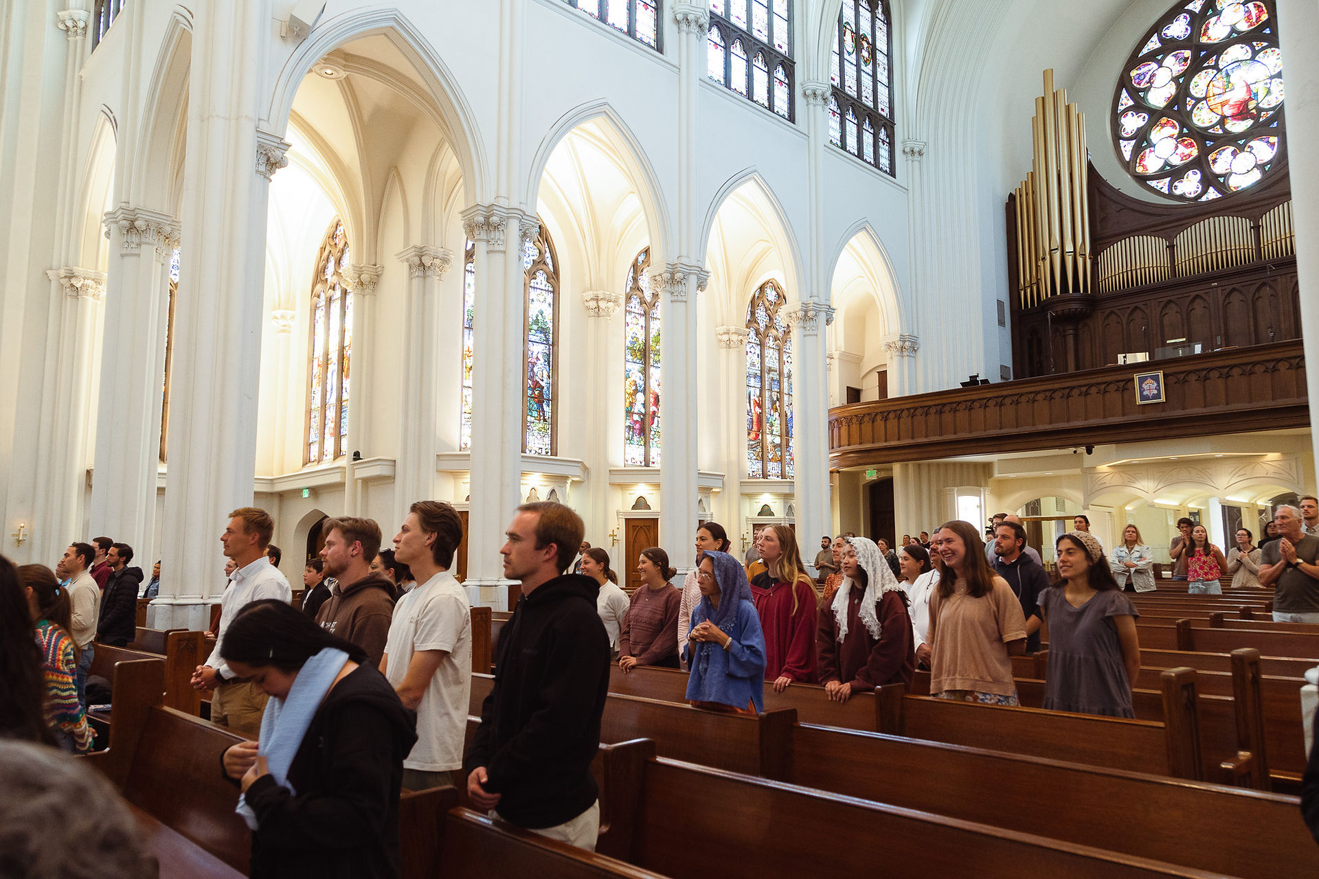 People stand in prayer inside a large church with stained glass windows and an organ. The atmosphere is serene and reverent.