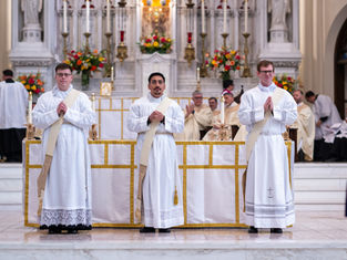 Three clergy in white robes stand before an ornate altar with candles and flowers, hands clasped in prayer, in a solemn church setting.