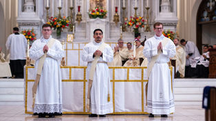 Three clergy in white robes stand before an ornate altar with candles and flowers, hands clasped in prayer, in a solemn church setting.