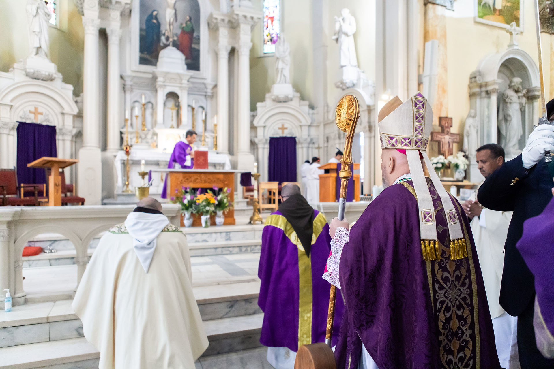 Bishops in purple robes stand in a church, one holding a staff, with altar and floral arrangements in the background. Stained glass visible.