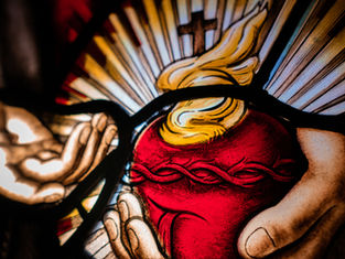 Stained glass art depicting hands holding a red heart with a crown of thorns, flames, and rays in the background, creating a luminous effect.