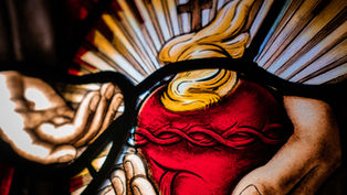 Stained glass art depicting hands holding a red heart with a crown of thorns, flames, and rays in the background, creating a luminous effect.