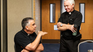 Two deacons in black clerical shirts talk in a meeting room, one seated gesturing, the other standing with a water bottle.