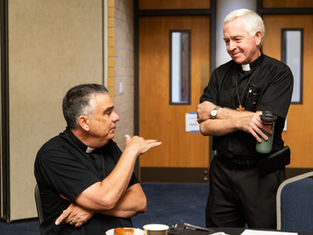 Two deacons in black clerical shirts talk in a meeting room, one seated gesturing, the other standing with a water bottle.