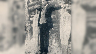 A man in a suit stands with arms outstretched, facing forward, next to ice sculptures in a snowy forest setting. Vintage black-and-white photo.