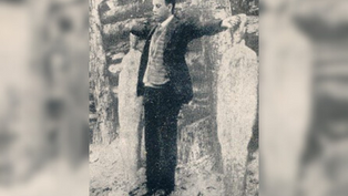 A man in a suit stands with arms outstretched, facing forward, next to ice sculptures in a snowy forest setting. Vintage black-and-white photo.