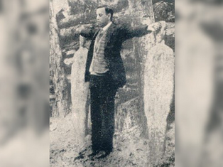 A man in a suit stands with arms outstretched, facing forward, next to ice sculptures in a snowy forest setting. Vintage black-and-white photo.