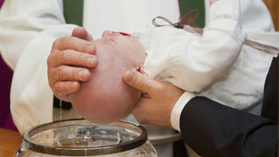 A baby in white is held over a crystal bowl during a baptism. A hand gently touches the baby's head, with water dripping down.