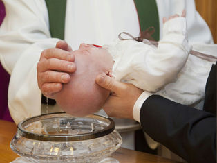 A baby in white is held over a crystal bowl during a baptism. A hand gently touches the baby's head, with water dripping down.