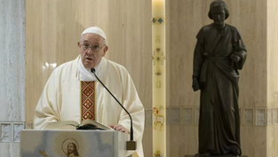 Man in white robe speaks at lectern with Bible; tall candle with symbols beside him; dark statue in background; solemn atmosphere.