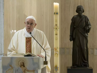 Man in white robe speaks at lectern with Bible; tall candle with symbols beside him; dark statue in background; solemn atmosphere.