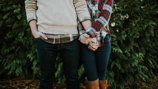 A couple holds hands, wearing casual fall attire with plaid and jeans. They're in front of green foliage, setting a cozy, intimate mood.