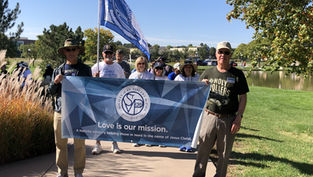Group of smiling adults hold a blue banner and flag in a sunny park; banner reads Love is our mission.