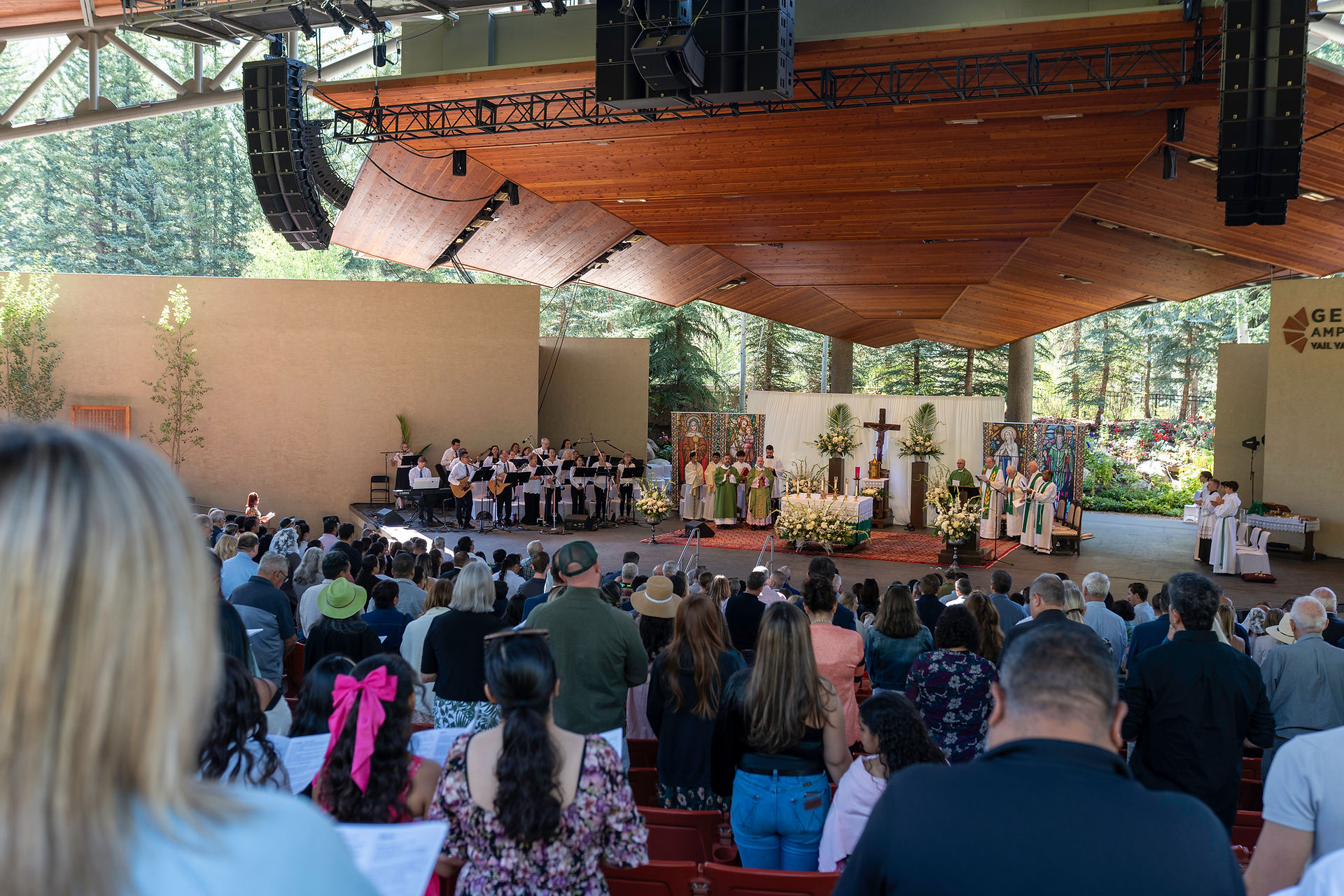 A large outdoor religious ceremony under a wooden pavilion, featuring an altar with a cross and floral arrangements. A choir stands to the left, and people in ceremonial attire flank the altar. Attendees are seated in red chairs, with trees and greenery in the background.