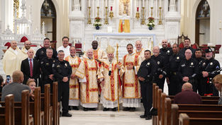 Clergy in ornate robes and police officers pose together in a decorated church with a white altar and gold accents, exuding a formal mood.