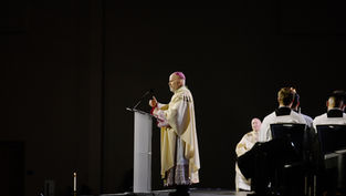 A clergyman in ornate robes speaks at a podium on a dimly lit stage, with seated attendees in white robes. Candle visible, somber mood.