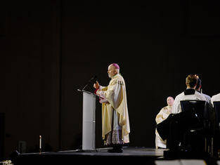 A clergyman in ornate robes speaks at a podium on a dimly lit stage, with seated attendees in white robes. Candle visible, somber mood.