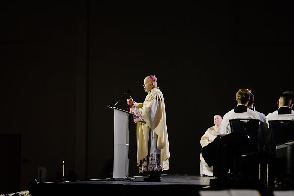 A clergyman in ornate robes speaks at a podium on a dimly lit stage, with seated attendees in white robes. Candle visible, somber mood.