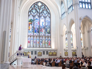 Priest in purple robe speaks from pulpit to seated congregation in bright cathedral with tall stained-glass windows
