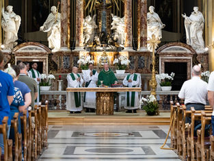 Catholic Mass in an ornate church, priests in green vestments at the altar while the congregation stands in pews before marble statues.