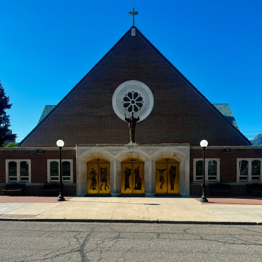 Brick church with a triangular roof, cross, and statue of Jesus on top. Golden doors with images below a circular window. Clear blue sky.