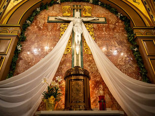Crucifix with white drapes displayed in a church, set against a marbled red wall. Floral arrangements at the base convey reverence.
