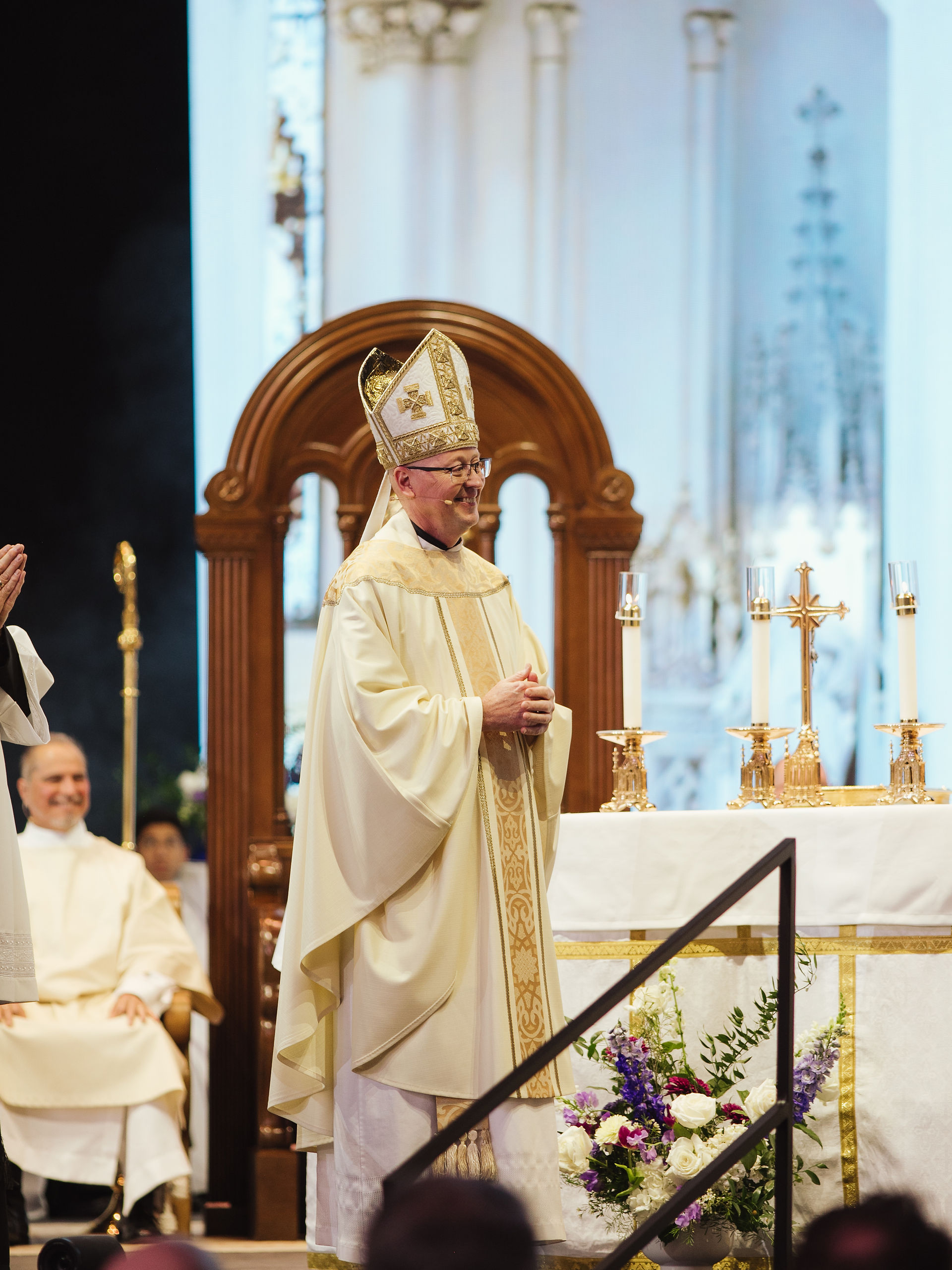 A bishop in elaborate robes stands at an altar, smiling during a ceremony. The setting is ornate with gold and floral accents.