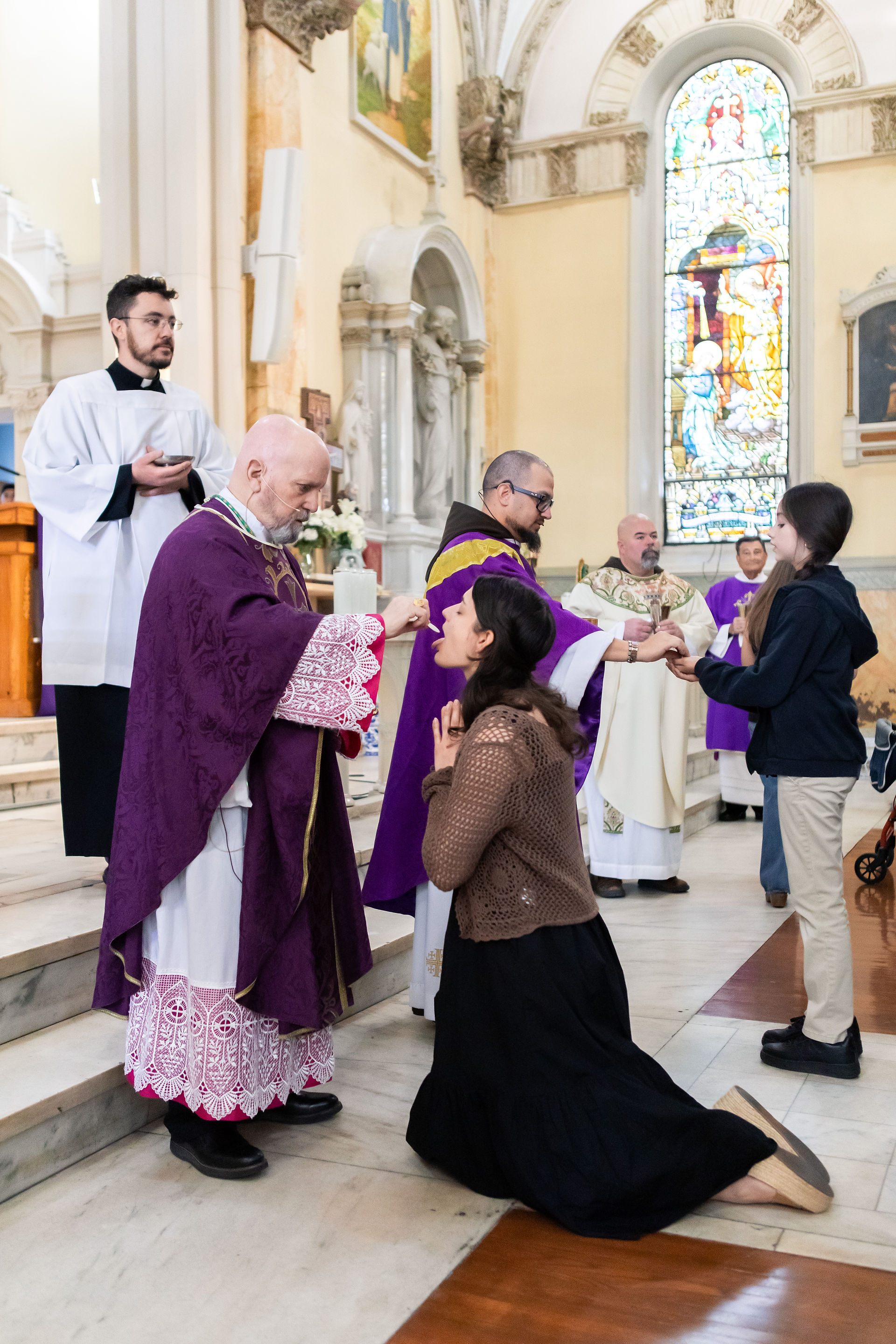 Clergy in purple and white vestments distribute communion to kneeling congregants inside a church with stained glass windows.