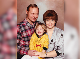 Family portrait: Smiling man in plaid shirt, woman in patterned blouse, and child in yellow dress with red bow, neutral background.