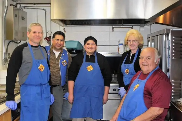Five smiling people in a kitchen wearing blue aprons with logos. Industrial background with stainless steel appliances. Warm, friendly mood.