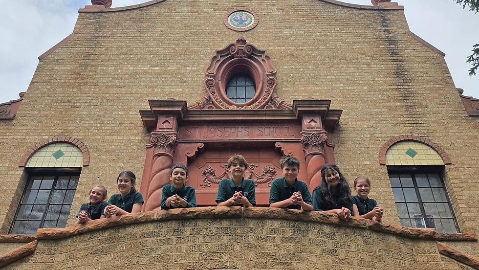 Smiling children in green uniforms lean on a balcony outside St. Joseph’s School, a brick historic building with ornate stonework.