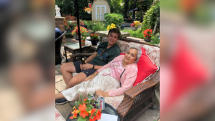 Young man and elderly woman relax on a wicker sofa in a garden with vibrant flowers and greenery, creating a warm and peaceful atmosphere.