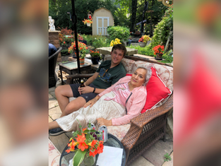 Young man and elderly woman relax on a wicker sofa in a garden with vibrant flowers and greenery, creating a warm and peaceful atmosphere.