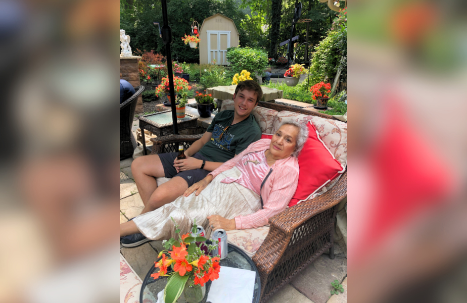 Young man and elderly woman relax on a wicker sofa in a garden with vibrant flowers and greenery, creating a warm and peaceful atmosphere.