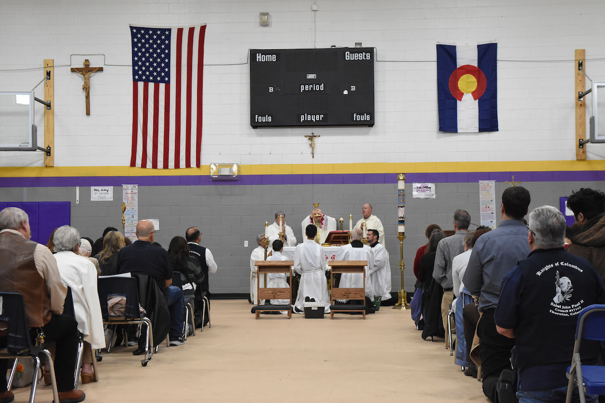A church service in a gym with a priest and altar servers in white robes. American and Colorado flags, plus a scoreboard, are visible.
