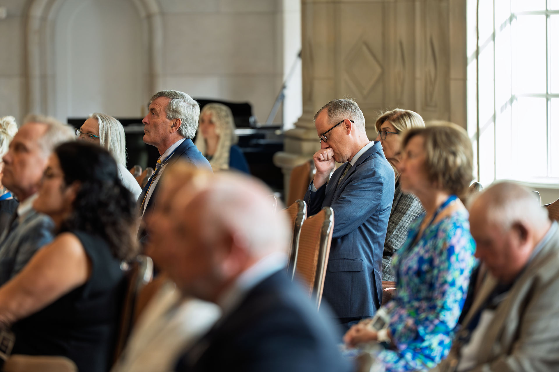 Audience seated, attentively listening in a formal setting with large windows. A man in a blue suit appears contemplative, room well-lit.