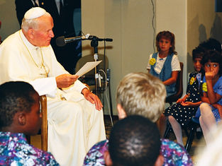 A person in white robes speaks into a mic, holding a paper. Children in colorful outfits listen attentively in a room with chairs.
