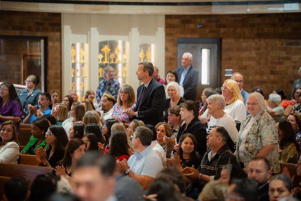 Congregants applaud in a church, with a man in a suit standing among seated attendees, warm wood interior.