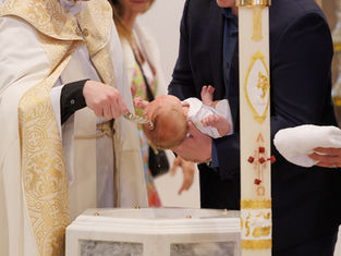 Priest pours water on a baby during a church baptism, with a man holding the infant beside a decorated paschal candle.