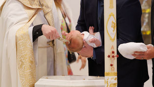 Priest pours water on a baby during a church baptism, with a man holding the infant beside a decorated paschal candle.