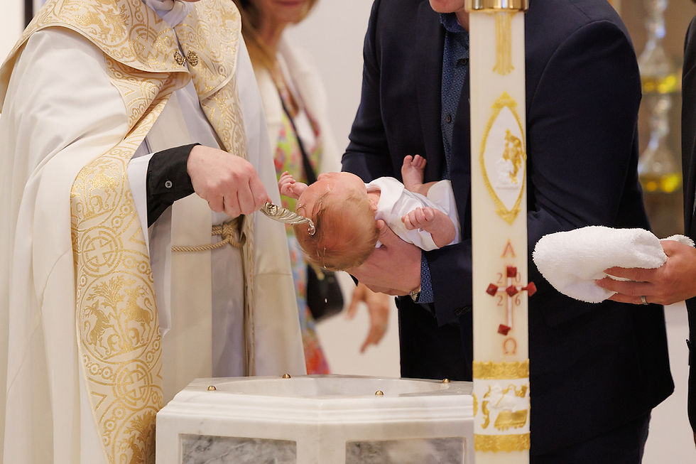 Priest pours water on a baby during a church baptism, with a man holding the infant beside a decorated paschal candle.